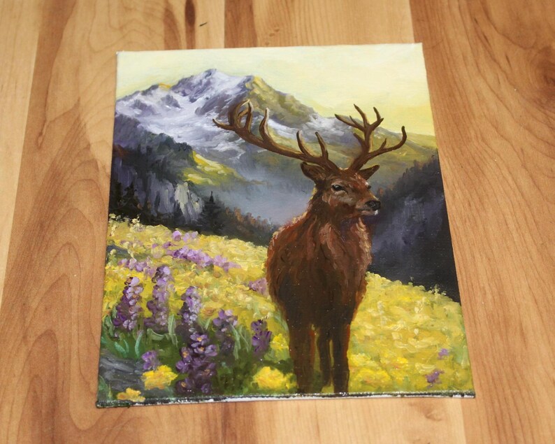 May include: An oil painting depicting a deer with large antlers in a field of yellow and purple wildflowers. The background shows a mountain range with snow-covered peaks and a soft, yellow sky. The deer is brown with a dark brown nose.