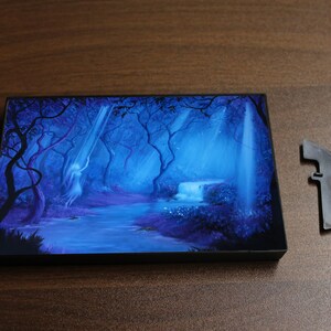 May include: A rectangular art piece with a fantasy forest scene in shades of blue and purple. A waterfall and a figure are visible. A detached black plastic piece is next to the art piece.