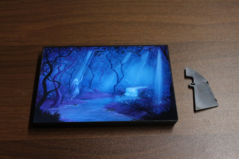 May include: A rectangular art piece with a fantasy forest scene in shades of blue and purple. A waterfall and a figure are visible. A detached black plastic piece is next to the art piece.