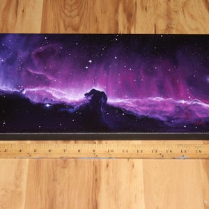 May include: A rectangular canvas print depicting a vibrant nebula in shades of purple, pink, and blue, with a prominent dark silhouette resembling a horse's head. The artwork is set against a starry background. A wooden ruler is placed below the canvas.