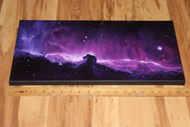 May include: A rectangular canvas print depicting a vibrant nebula in shades of purple, pink, and blue, with a prominent dark silhouette resembling a horse's head. The artwork is set against a starry background. A wooden ruler is placed below the canvas.