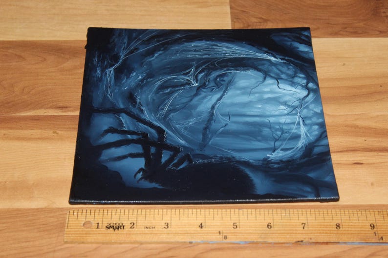 May include: A square canvas painting featuring a dark blue and black forest scene with a large spider silhouette in the foreground. A wooden ruler is placed along the bottom edge, showing the canvas width is approximately 9 inches wide.