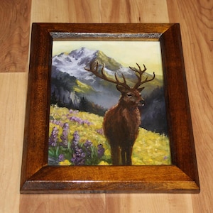 May include: A framed painting depicting a deer in a mountain scene. The artwork shows a brown deer with large antlers in a field of yellow and purple flowers. The background features a mountain range with snow-covered peaks.
