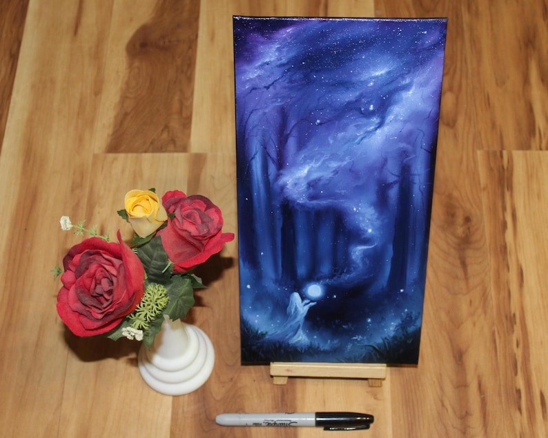 May include: A rectangular canvas painting depicting a dark forest scene with a glowing orb held by a figure, set on a small wooden easel. Beside it is a white vase containing red and yellow roses, with a black permanent marker measuring 5.25 inches.