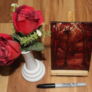 K&ouml;nnte beinhalten: Ein Stillleben mit zwei roten Rosen in einer wei&szlig;en Vase, einem kleinen Gem&auml;lde einer Waldszene und einem schwarzen und silbernen Sharpie-Marker. Das Gem&auml;lde befindet sich auf einer Holzstaffelei.