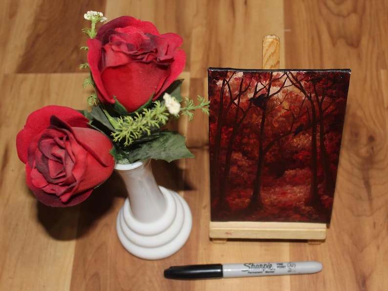 K&ouml;nnte beinhalten: Ein Stillleben mit zwei roten Rosen in einer wei&szlig;en Vase, einem kleinen Gem&auml;lde einer Waldszene und einem schwarzen und silbernen Sharpie-Marker. Das Gem&auml;lde befindet sich auf einer Holzstaffelei.