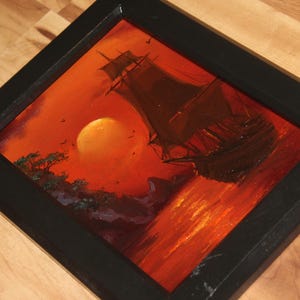 Pu&ograve; includere: Un dipinto incorniciato che raffigura un veliero sull'acqua durante un tramonto. La scena presenta un grande sole arancione, sagome scure di una nave e di una costa rocciosa, e vivaci tonalit&agrave; di rosso e arancione sull'acqua.