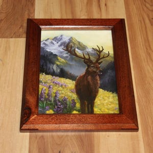 May include: A framed painting depicting a deer in a mountain scene. The artwork shows a brown deer with large antlers in a field of yellow and purple flowers, with mountains in the background. The frame is made of dark wood.