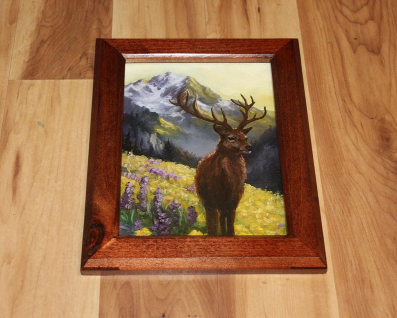 May include: A framed painting depicting a deer in a mountain scene. The artwork shows a brown deer with large antlers in a field of yellow and purple flowers, with mountains in the background. The frame is made of dark wood.