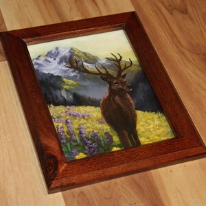 May include: A framed painting depicting a deer in a field of yellow and purple flowers, with mountains in the background. The frame is made of dark brown wood.
