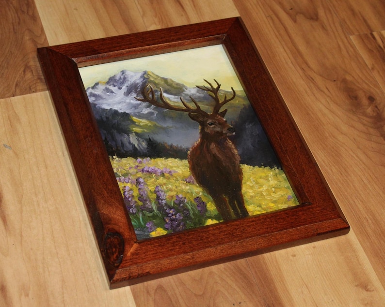May include: A framed painting depicting a deer in a field of yellow and purple flowers, with mountains in the background. The frame is made of dark brown wood.