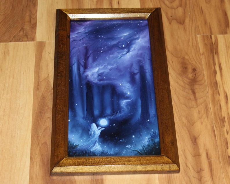 May include: A rectangular framed print featuring a dark blue forest scene at night. A figure in a white robe stands in the foreground holding a glowing orb. Swirling blue mist rises from the orb into the trees under a starry night sky.