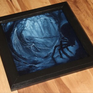 May include: A square wall art piece featuring a dark, moody illustration of a spider in a web-filled forest. The artwork is rendered in shades of deep blue and black, encased in a simple black frame, depicting a forest scene with a large spider.