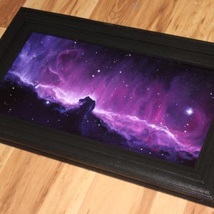 May include: A framed artwork depicting a vibrant nebula in shades of purple, pink, and blue, with numerous stars. The artwork is encased in a black rectangular frame, creating a striking contrast. The piece is displayed on a wooden surface.