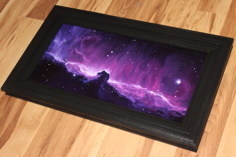 May include: A framed artwork depicting a vibrant nebula in shades of purple, pink, and blue, with numerous stars. The artwork is encased in a black rectangular frame, creating a striking contrast. The piece is displayed on a wooden surface.