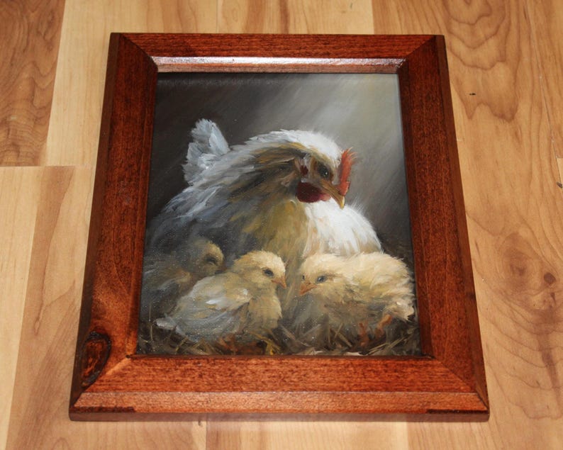 May include: A framed painting depicts a hen with three chicks. The hen is white with gray shading, a red comb, and a yellow beak. The chicks are yellow and fluffy. The frame is a dark brown wood.