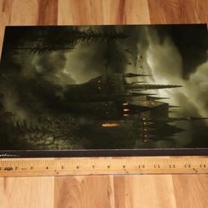 May include: A canvas print depicting a gothic castle under a stormy sky. The artwork features a dark color palette with shades of green and gray, and the castle is illuminated by warm, golden lights. The print is rectangular and rests on a wooden surface.