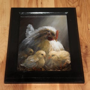 May include: A framed painting depicts a hen with three chicks. The hen has white and gray feathers, a red comb, and a yellow beak. The chicks are yellow and fluffy. The painting is in a black frame and is set against a dark background.