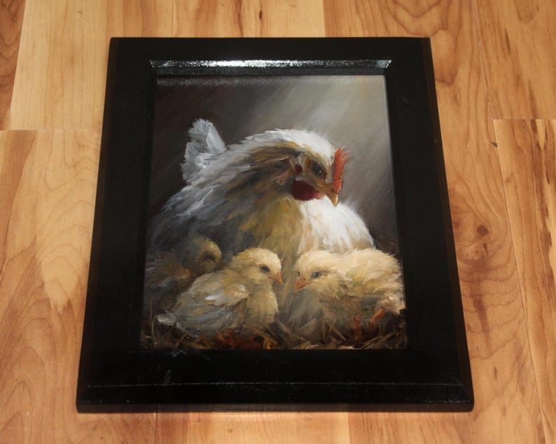 May include: A framed painting depicts a hen with three chicks. The hen has white and gray feathers, a red comb, and a yellow beak. The chicks are yellow and fluffy. The painting is in a black frame and is set against a dark background.