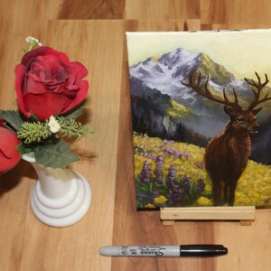 May include: An oil painting of a deer with large antlers in a mountain landscape, displayed on a small wooden easel. Beside it, a small white vase holds two red roses. A black Sharpie marker rests on the wooden surface.