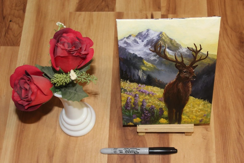 May include: An oil painting of a deer with large antlers in a mountain landscape, displayed on a small wooden easel. Beside it, a small white vase holds two red roses. A black Sharpie marker rests on the wooden surface.