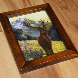May include: A framed painting of a deer in a field of yellow and purple flowers, with a mountain range in the background. The frame is made of dark brown wood. The artwork depicts a majestic deer with large antlers.