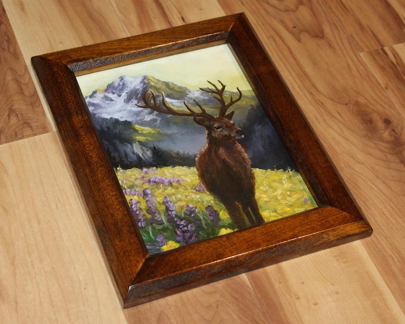 May include: A framed painting of a deer in a field of yellow and purple flowers, with a mountain range in the background. The frame is made of dark brown wood. The artwork depicts a majestic deer with large antlers.
