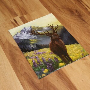 May include: An oil painting depicting a deer with antlers in a field of yellow and purple flowers. Mountains are in the background with a yellow and light blue sky. The painting is on a square canvas.