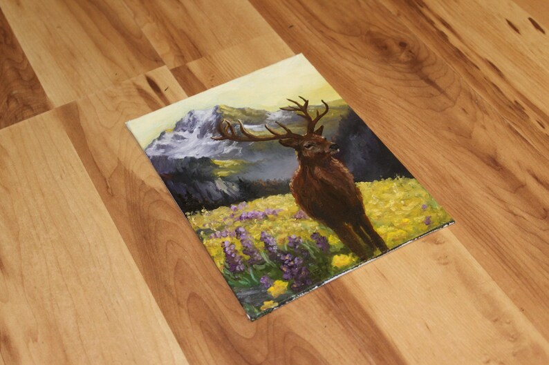 May include: An oil painting depicting a deer with antlers in a field of yellow and purple flowers. Mountains are in the background with a yellow and light blue sky. The painting is on a square canvas.