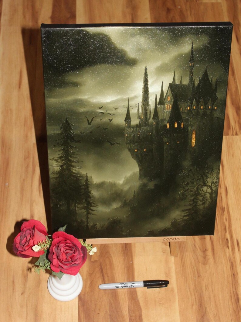 May include: A canvas print of a gothic castle at night, with a dark, moody color palette. The artwork features a detailed depiction of the castle with multiple spires, set against a cloudy sky. A small vase of red roses and a black marker are in the foreground.