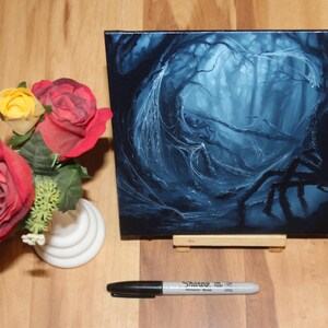 May include: A square canvas painting depicting a dark, blue-toned forest scene with a large spider silhouette, displayed on a small wooden easel. Beside it sits a white vase with red and yellow artificial roses and a black Sharpie marker.