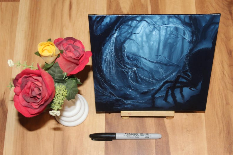 May include: A square canvas painting depicting a dark, blue-toned forest scene with a large spider silhouette, displayed on a small wooden easel. Beside it sits a white vase with red and yellow artificial roses and a black Sharpie marker.
