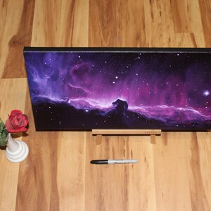 May include: A rectangular canvas print depicting a vibrant nebula in shades of purple and pink, with a black frame. The print is displayed on a wooden stand. A small white vase with red roses and a black marker are also present.