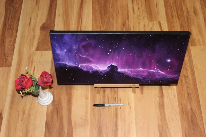 May include: A rectangular canvas print depicting a vibrant nebula in shades of purple and pink, with a black frame. The print is displayed on a wooden stand. A small white vase with red roses and a black marker are also present.