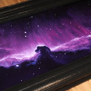 May include: A framed painting of a nebula in shades of purple, pink, and blue. The artwork depicts a cosmic scene with swirling clouds of gas and dust, and scattered stars. The black frame adds a contrasting border to the vibrant space art.