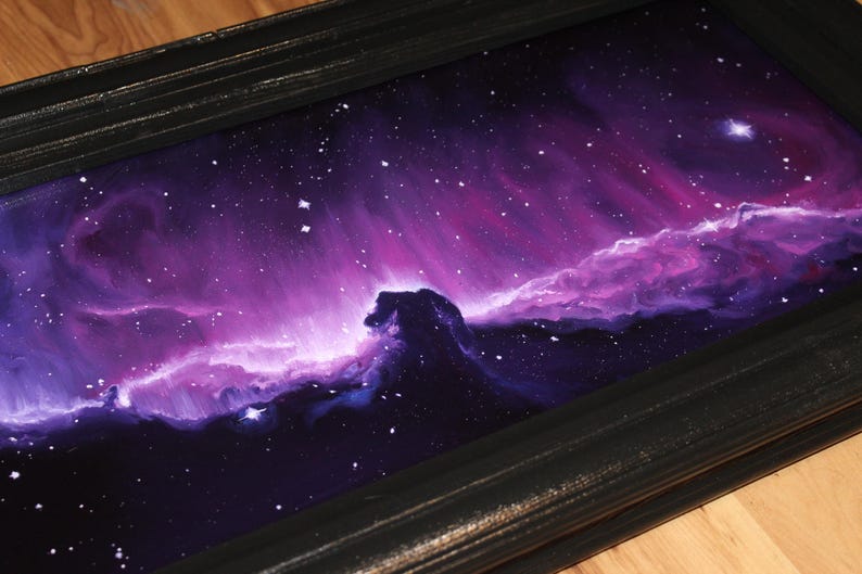 May include: A framed painting of a nebula in shades of purple, pink, and blue. The artwork depicts a cosmic scene with swirling clouds of gas and dust, and scattered stars. The black frame adds a contrasting border to the vibrant space art.