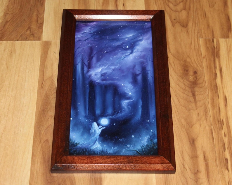 May include: A rectangular framed print featuring a dark blue forest scene with a glowing white orb held by a figure in a flowing garment. The artwork is encased in a polished dark brown wooden frame with a smooth, glossy finish and beveled edges.