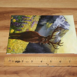 May include: An original painting of a deer with large antlers in a natural landscape. The artwork features a brown deer against a backdrop of yellow and purple flowers, a waterfall, and a mountain. The canvas measures approximately 20 cm in length.