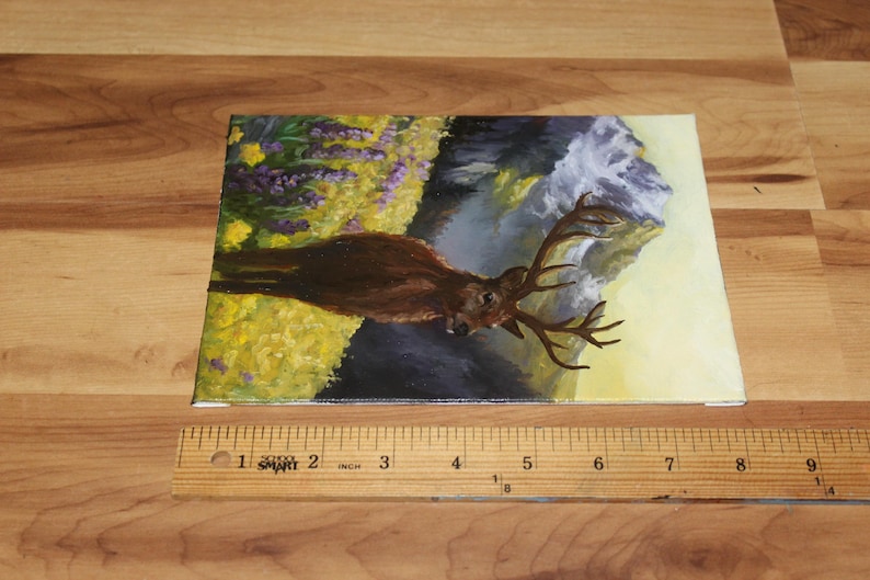 May include: An original painting of a deer with large antlers in a natural landscape. The artwork features a brown deer against a backdrop of yellow and purple flowers, a waterfall, and a mountain. The canvas measures approximately 20 cm in length.