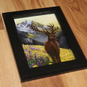 May include: A framed painting depicting a deer in a mountain scene. The artwork shows a brown deer with antlers in a field of yellow and purple flowers, with mountains in the background. The frame is black.