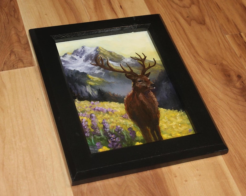 May include: A framed painting depicting a deer in a mountain scene. The artwork shows a brown deer with antlers in a field of yellow and purple flowers, with mountains in the background. The frame is black.