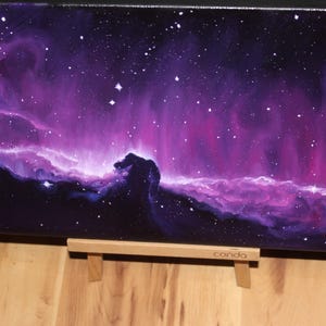 May include: A canvas painting depicting a vibrant nebula in shades of purple, pink, and blue, with numerous white stars scattered across the dark background. The painting is displayed on a wooden easel. The word "conda" is visible on the easel.
