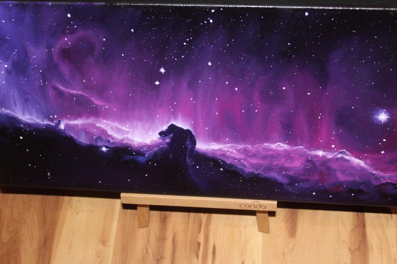 May include: A canvas painting depicting a vibrant nebula in shades of purple, pink, and blue, with numerous white stars scattered across the dark background. The painting is displayed on a wooden easel. The word "conda" is visible on the easel.