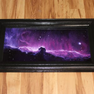 May include: A rectangular black framed art piece featuring a vibrant depiction of a nebula in shades of purple, pink, and white. The artwork showcases a celestial scene with stars and cosmic clouds, creating a dramatic and colorful display.