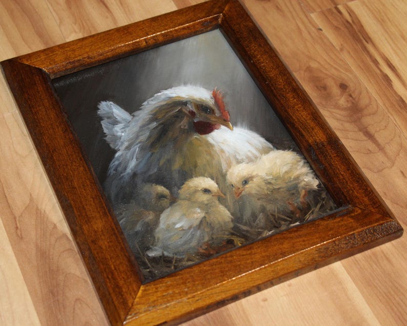 May include: A framed painting depicts a hen with three chicks. The hen is white and gray with a red comb. The chicks are yellow and fluffy. The frame is a dark brown wood.