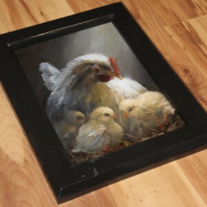 May include: A framed painting of a hen with three chicks. The hen is white with a red comb, and the chicks are yellow. The painting is in a black frame and is set against a dark background.
