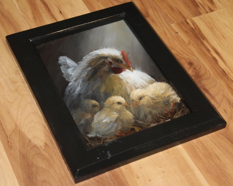 May include: A framed painting of a hen with three chicks. The hen is white with a red comb, and the chicks are yellow. The painting is in a black frame and is set against a dark background.
