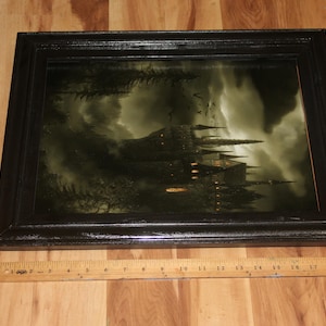 May include: A framed print depicting a gothic castle under a stormy sky. The castle has multiple spires and lit windows. The frame is black and rectangular. A wooden ruler is placed below the frame.
