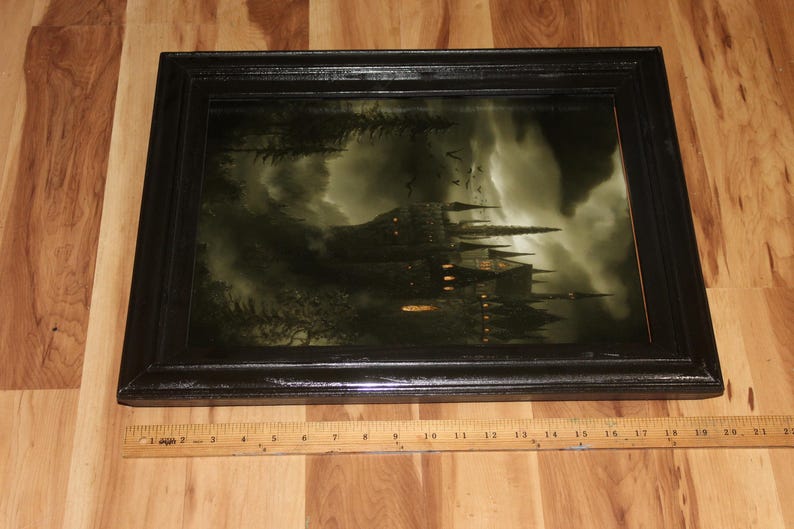 May include: A framed print depicting a gothic castle under a stormy sky. The castle has multiple spires and lit windows. The frame is black and rectangular. A wooden ruler is placed below the frame.