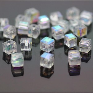 Könnte beinhalten: Eine Ansammlung klarer, irisierender, würfelförmiger Perlen. Die Perlen haben eine facettierte Oberfläche, die das Licht reflektiert und einen Regenbogeneffekt erzeugt. Jede Perle hat ein Loch in der Mitte, was darauf hindeutet, dass sie zum Auffädeln gedacht sind.